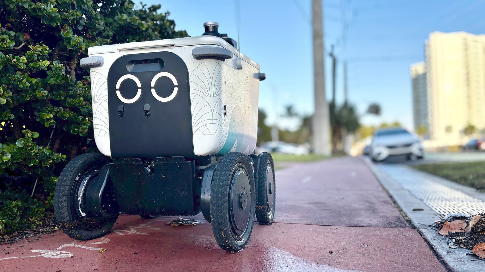 A small delivery robot stopped on a city sidewalk near a pedestrian crossing