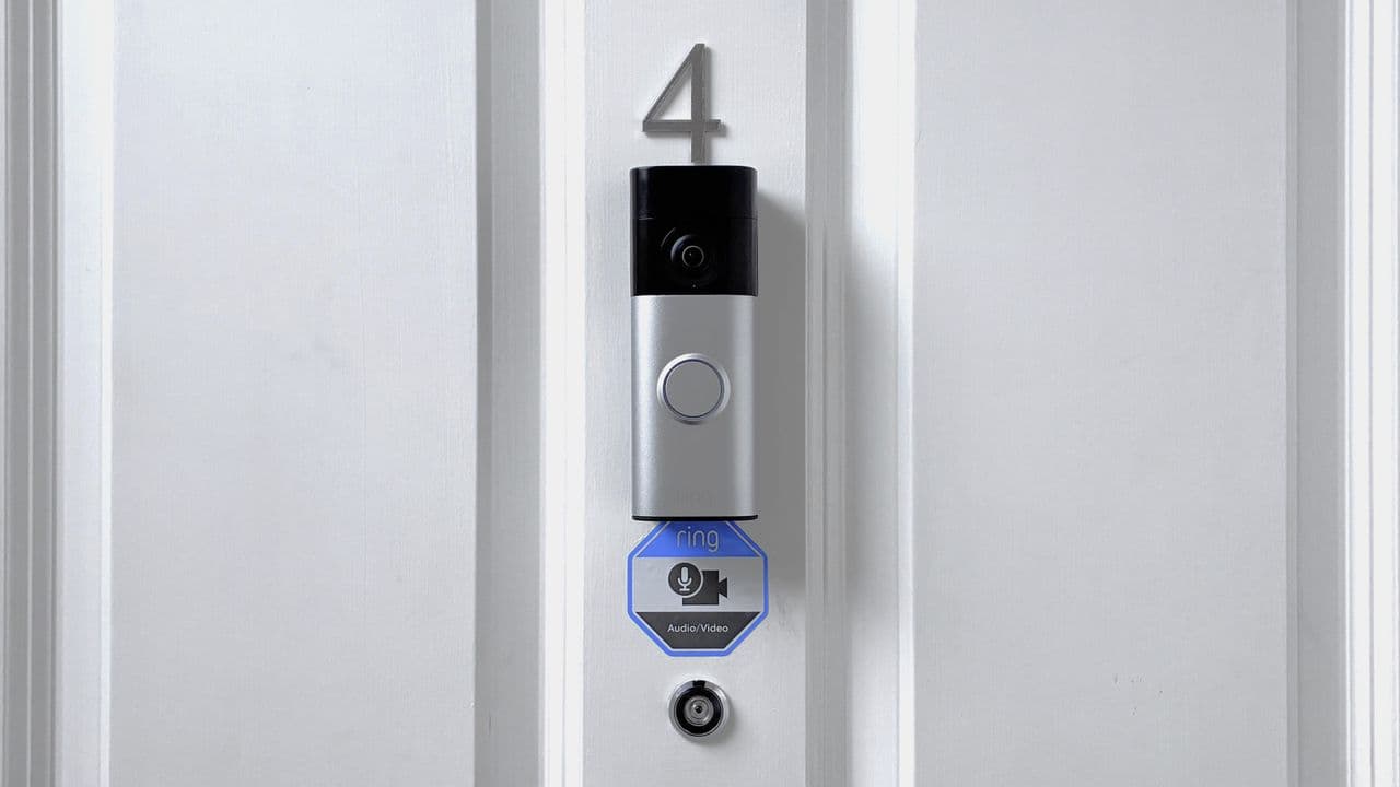 A white Ring Battery Video Doorbell Plus mounted beside a wooden front door showing its slim profile