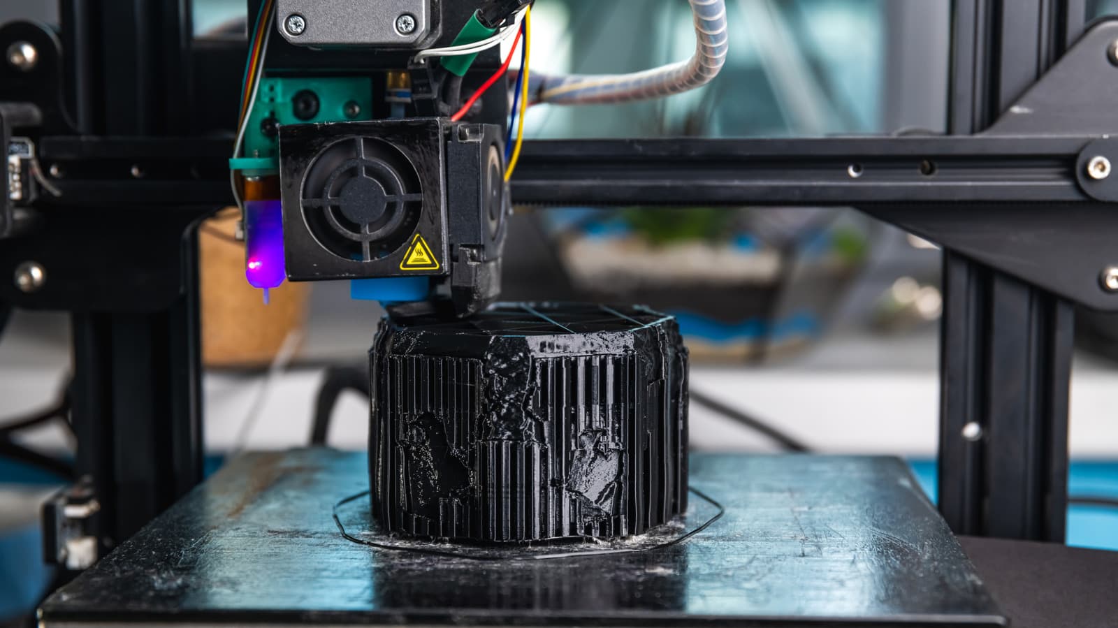 A 3D printer in operation with visible print bed and extruder, showing signs of use and wear.