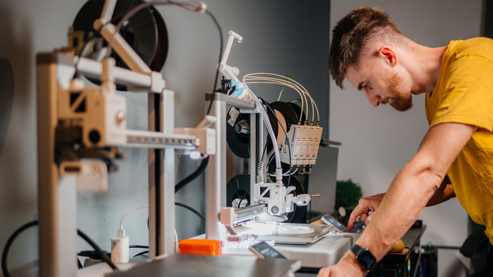 A desktop 3D printer with a partially completed plastic model on its build plate, surrounded by filament spools and tools