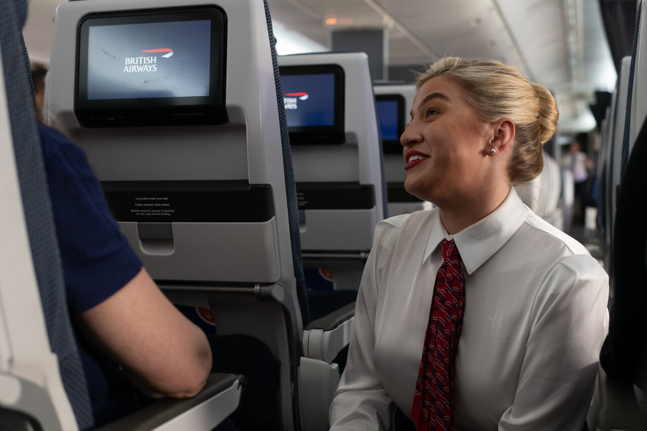 A passenger wearing Meta Ray-Ban smart glasses on a British Airways flight, with a cabin crew member in the background